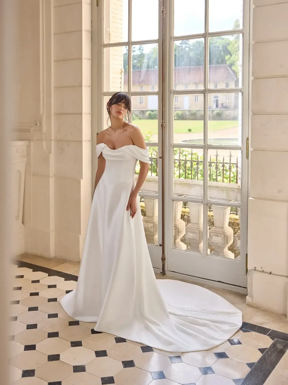 A woman in a white off-the-shoulder wedding dress standing near a large window with a scenic view.