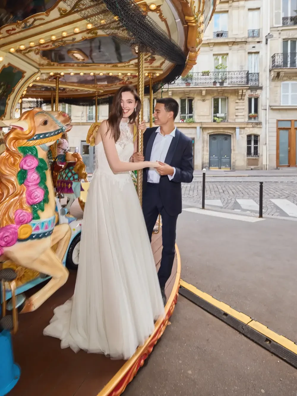 A couple stands together on a carousel, with the woman in a flowing white wedding dress and the man in a dark suit, surrounded by a picturesque street scene.